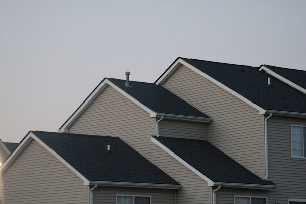Several suburban houses with gabled roofs are aligned in a row under a gray sky. The houses are uniformly built with beige siding and black asphalt shingle roofs, giving a uniform and orderly appearance.