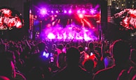 A vibrant concert scene at night with a large crowd gathered in front of a brightly lit stage. There are colorful lights illuminating the area, and people in the audience are raising their hands, holding up phones, and some are wearing illuminated headbands.
