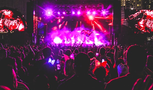 A vibrant concert scene at night with a large crowd gathered in front of a brightly lit stage. There are colorful lights illuminating the area, and people in the audience are raising their hands, holding up phones, and some are wearing illuminated headbands.