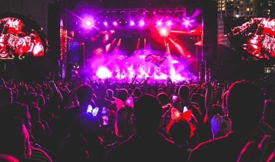 A vibrant concert scene at night with a large crowd gathered in front of a brightly lit stage. There are colorful lights illuminating the area, and people in the audience are raising their hands, holding up phones, and some are wearing illuminated headbands.
