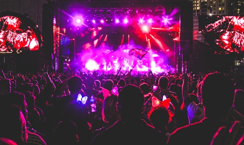 A vibrant concert scene at night with a large crowd gathered in front of a brightly lit stage. There are colorful lights illuminating the area, and people in the audience are raising their hands, holding up phones, and some are wearing illuminated headbands.