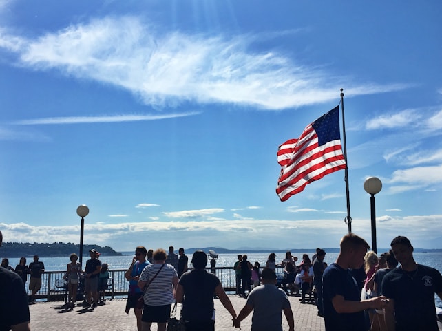A group of people gather near a waterfront under a clear blue sky with scattered clouds. An American flag waves prominently in the wind, mounted on a pole. The scene includes people walking and chatting, with a backdrop of a distant shoreline and water, creating a lively outdoor atmosphere.