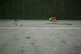 A person sits alone on an empty court with knees drawn to their chest, head resting on their knees. The court surface is gray with cracks and stains, and a green wall is in the background. There is a tennis racket laying on the ground nearby, as well as some scattered debris.