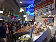 A busy indoor fish market with a variety of seafood on display, including large crabs and fish. The market is crowded with people, some browsing and some engaging with the sellers. There are multiple signs advertising the fresh seafood, including neon signs and banners. The lighting is bright, with several overhead lamps illuminating the market area.