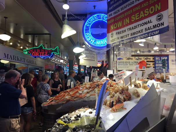 A busy indoor fish market with a variety of seafood on display, including large crabs and fish. The market is crowded with people, some browsing and some engaging with the sellers. There are multiple signs advertising the fresh seafood, including neon signs and banners. The lighting is bright, with several overhead lamps illuminating the market area.