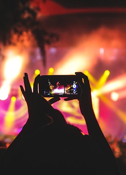 person holding smartphone taking video of a concert near stage with lights during nighttime