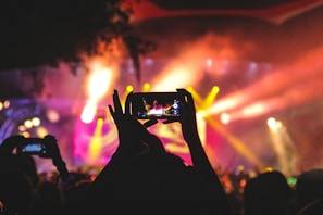 A vibrant live concert scene is captured with colorful stage lights creating an energetic atmosphere. The silhouette of a person holding a smartphone is visible in the foreground, as they take photos or videos of the performance.