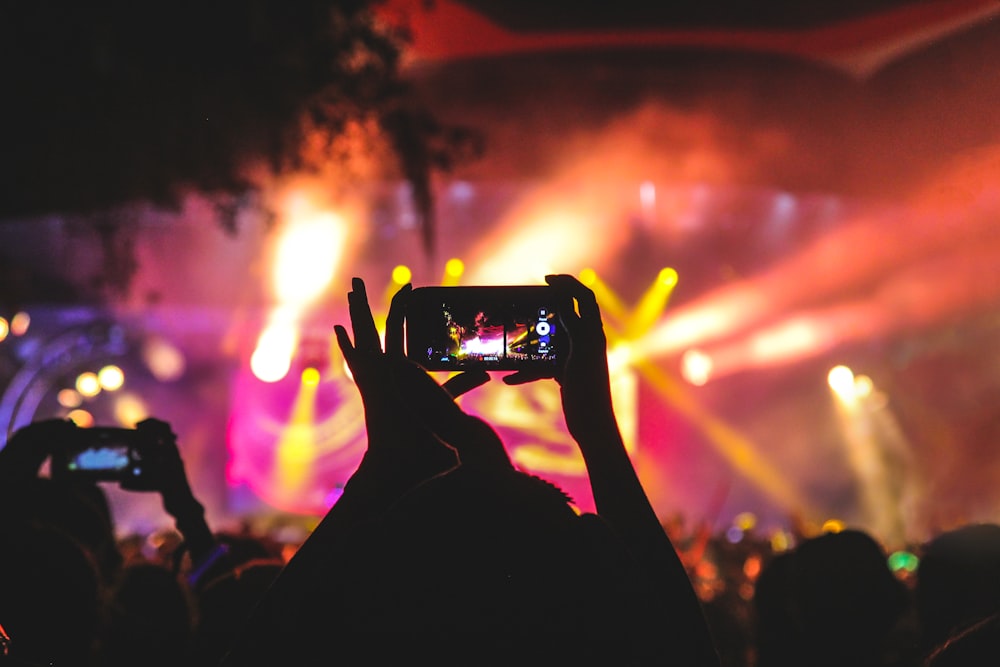 person holding smartphone taking video of a concert near stage with lights during nighttime