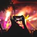 person holding smartphone taking video of a concert near stage with lights during nighttime