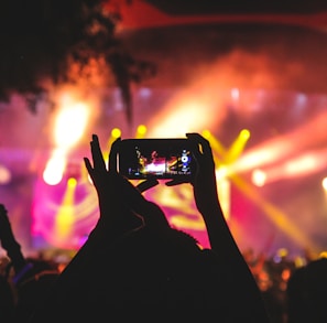 person holding smartphone taking video of a concert near stage with lights during nighttime