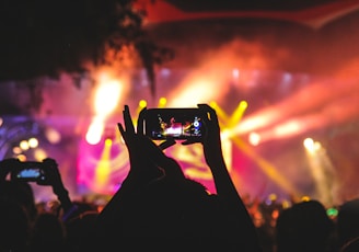 person holding smartphone taking video of a concert near stage with lights during nighttime