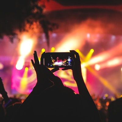 person holding smartphone taking video of a concert near stage with lights during nighttime