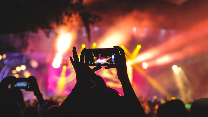 person holding smartphone taking video of a concert near stage with lights during nighttime