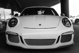 A sleek sports car with a white exterior, featuring modern, rounded headlights and a prominent front grille with mesh detailing. The car's streamlined design and low profile exemplify luxury and performance. The setting appears to be a showroom or dealership, indicated by the reflective glass and other vehicles in the background.