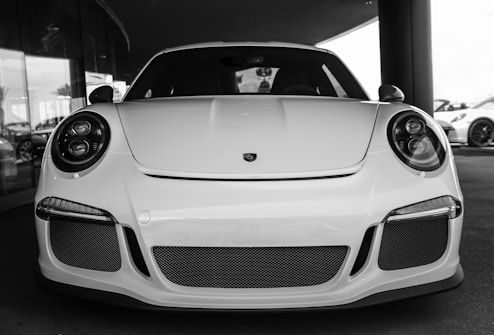 A sleek sports car with a white exterior, featuring modern, rounded headlights and a prominent front grille with mesh detailing. The car's streamlined design and low profile exemplify luxury and performance. The setting appears to be a showroom or dealership, indicated by the reflective glass and other vehicles in the background.