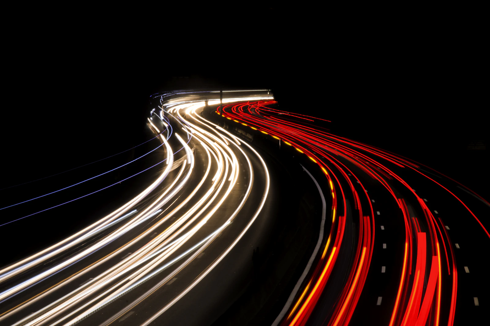 Long-exposure white and red car light trails sweeping around a dark highway curve at night
