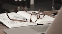 An open notebook with handwritten notes is placed on a table, accompanied by a pair of reading glasses and a pen. The scene features a laptop and a blurred background, suggesting an office or study environment.
