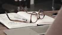 An open notebook with handwritten notes is placed on a table, accompanied by a pair of reading glasses and a pen. The scene features a laptop and a blurred background, suggesting an office or study environment.