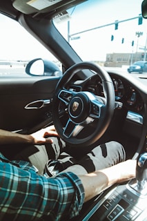 A person is seated inside a luxury car, holding the steering wheel with one hand while the other hand rests near the gear shift. The interior features a modern dashboard with a visible brand logo in the center of the steering wheel. Sunlight streams through the window, highlighting the plaid shirt of the driver. Outside, a road and traffic lights are visible through the windshield.