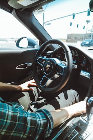 A person is seated inside a luxury car, holding the steering wheel with one hand while the other hand rests near the gear shift. The interior features a modern dashboard with a visible brand logo in the center of the steering wheel. Sunlight streams through the window, highlighting the plaid shirt of the driver. Outside, a road and traffic lights are visible through the windshield.