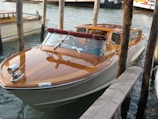 A sleek wooden motorboat with a polished finish is moored between wooden posts in a waterway. The boat has a glossy deck with a maroon cushion on its windshield, set against a backdrop of calm water.