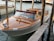 A sleek wooden motorboat with a polished finish is moored between wooden posts in a waterway. The boat has a glossy deck with a maroon cushion on its windshield, set against a backdrop of calm water.