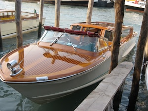 A sleek wooden motorboat with a polished finish is moored between wooden posts in a waterway. The boat has a glossy deck with a maroon cushion on its windshield, set against a backdrop of calm water.
