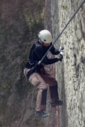 A person equipped with a helmet and climbing gear is scaling a textured vertical wall outdoors. The climber is wearing a black long-sleeve shirt, brown pants, and sturdy climbing shoes. The scene includes safety ropes and various climbing holds attached to the wall.