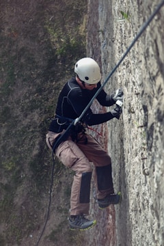 A person equipped with a helmet and climbing gear is scaling a textured vertical wall outdoors. The climber is wearing a black long-sleeve shirt, brown pants, and sturdy climbing shoes. The scene includes safety ropes and various climbing holds attached to the wall.