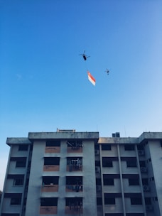 A residential building stands tall under a clear blue sky. Two helicopters are flying above, carrying a large flag with red and white stripes.