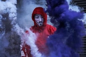 A person wearing a red hoodie and a Guy Fawkes mask is surrounded by thick clouds of blue and white smoke. The background is obscured, adding to the mysterious and dramatic atmosphere.