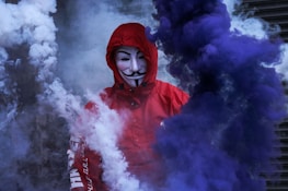A person wearing a red hoodie and a Guy Fawkes mask is surrounded by thick clouds of blue and white smoke. The background is obscured, adding to the mysterious and dramatic atmosphere.