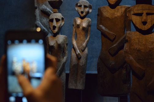 A collection of large, carved wooden sculptures depicting stylized human figures is observed through a smartphone camera. The figures appear abstract and simplistic, with angular features, set against a dimly lit background that emphasizes their texture.