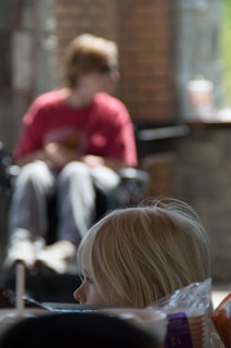 A young child with blond hair appears in the foreground, looking to the side with a thoughtful expression. In the background, a figure wearing a red shirt and sunglasses sits in a wheelchair, slightly out of focus. The setting includes a mix of natural and artificial lighting, with brick walls and some blurred objects, possibly creating a cozy atmosphere.