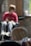 A young child with blond hair appears in the foreground, looking to the side with a thoughtful expression. In the background, a figure wearing a red shirt and sunglasses sits in a wheelchair, slightly out of focus. The setting includes a mix of natural and artificial lighting, with brick walls and some blurred objects, possibly creating a cozy atmosphere.