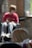 A young child with blond hair appears in the foreground, looking to the side with a thoughtful expression. In the background, a figure wearing a red shirt and sunglasses sits in a wheelchair, slightly out of focus. The setting includes a mix of natural and artificial lighting, with brick walls and some blurred objects, possibly creating a cozy atmosphere.