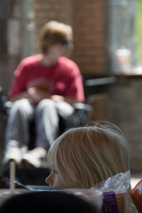 A young child with blond hair appears in the foreground, looking to the side with a thoughtful expression. In the background, a figure wearing a red shirt and sunglasses sits in a wheelchair, slightly out of focus. The setting includes a mix of natural and artificial lighting, with brick walls and some blurred objects, possibly creating a cozy atmosphere.
