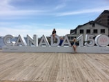 A large sign displaying 'CANADA 150' is placed on a wooden dock with three people sitting on different letters of the sign. The background features a body of water, part of a cityscape with buildings, and a clear sky with scattered clouds. One of the buildings has the word 'SALTY'S' visible.