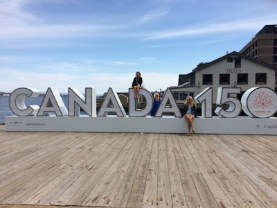 A large sign displaying 'CANADA 150' is placed on a wooden dock with three people sitting on different letters of the sign. The background features a body of water, part of a cityscape with buildings, and a clear sky with scattered clouds. One of the buildings has the word 'SALTY'S' visible.