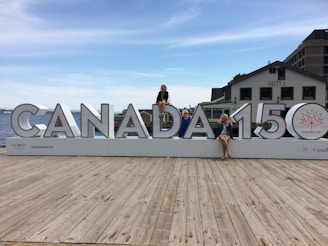A large sign displaying 'CANADA 150' is placed on a wooden dock with three people sitting on different letters of the sign. The background features a body of water, part of a cityscape with buildings, and a clear sky with scattered clouds. One of the buildings has the word 'SALTY'S' visible.