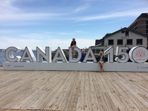 A large sign displaying 'CANADA 150' is placed on a wooden dock with three people sitting on different letters of the sign. The background features a body of water, part of a cityscape with buildings, and a clear sky with scattered clouds. One of the buildings has the word 'SALTY'S' visible.