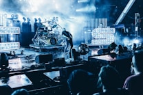 A live concert scene features a vibrant stage with intense lighting and fog effects. Visible are large illuminated letters, a musician playing guitar, and a drum set. The audience is partially visible in the foreground, creating an energetic atmosphere.