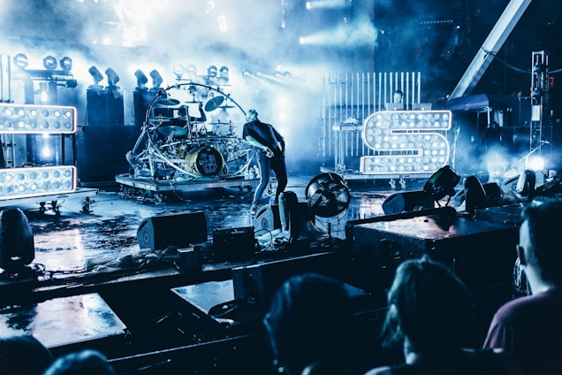 A live concert scene features a vibrant stage with intense lighting and fog effects. Visible are large illuminated letters, a musician playing guitar, and a drum set. The audience is partially visible in the foreground, creating an energetic atmosphere.