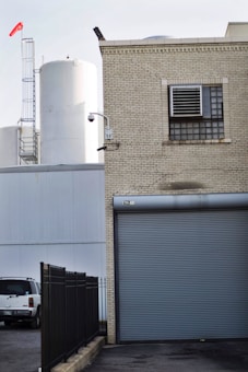 A brick industrial building with a roll-up door and ventilation system is situated adjacent to large white storage tanks. A security camera is mounted on the corner of the building. A black fence runs parallel to the building, and a white vehicle is parked nearby. A red windsock is visible atop a ladder structure.