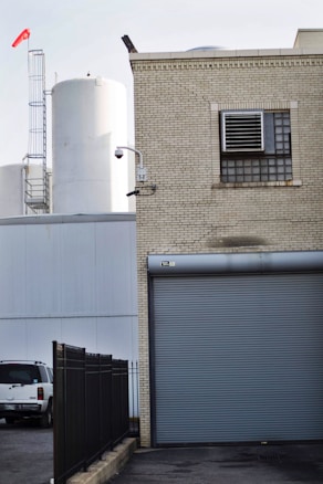 A brick industrial building with a roll-up door and ventilation system is situated adjacent to large white storage tanks. A security camera is mounted on the corner of the building. A black fence runs parallel to the building, and a white vehicle is parked nearby. A red windsock is visible atop a ladder structure.