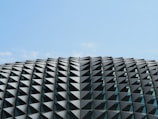 A modern architectural structure featuring a dome-like shape composed of numerous triangular panels. The panels create a geometric pattern and are made of reflective material, adding a sleek and contemporary look.