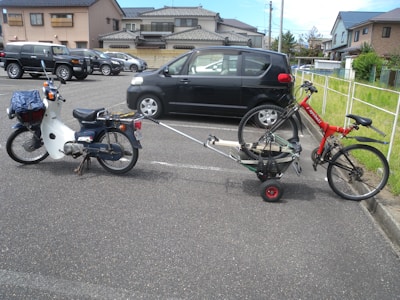 A motorbike is parked in a residential area with a bicycle attached to it via a makeshift trailer. The trailer is constructed with a small platform on wheels. The scene includes parked cars and residential houses in the background.