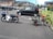 A motorbike is parked in a residential area with a bicycle attached to it via a makeshift trailer. The trailer is constructed with a small platform on wheels. The scene includes parked cars and residential houses in the background.