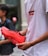 A person holding a vibrant orange sports shoe with translucent soles, standing next to someone else. The person is wearing a white t-shirt with a distinct logo in red and black. The background is muted, suggesting an urban environment.