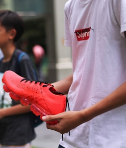 A person holding a vibrant orange sports shoe with translucent soles, standing next to someone else. The person is wearing a white t-shirt with a distinct logo in red and black. The background is muted, suggesting an urban environment.